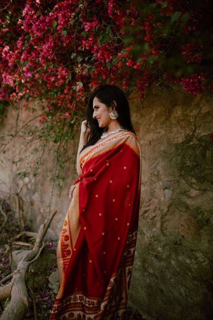 Positive young Indian lady in red traditional clothes standing near stone hill with green plants with flowers in daylight in nature