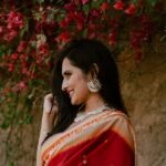 Positive young Indian lady in red traditional clothes standing near stone hill with green plants with flowers in daylight in nature