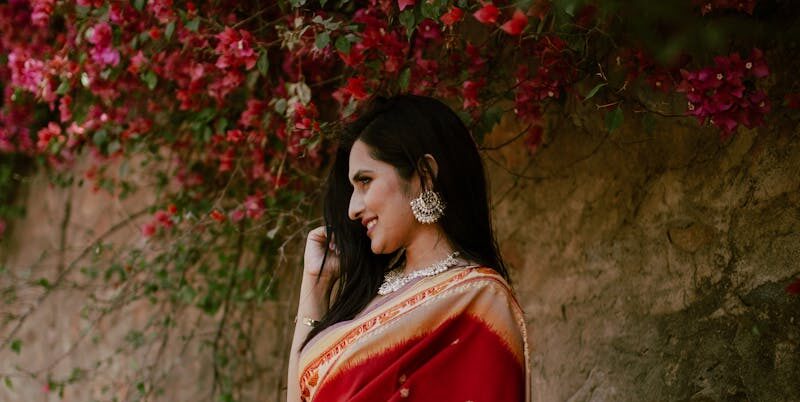 Positive young Indian lady in red traditional clothes standing near stone hill with green plants with flowers in daylight in nature